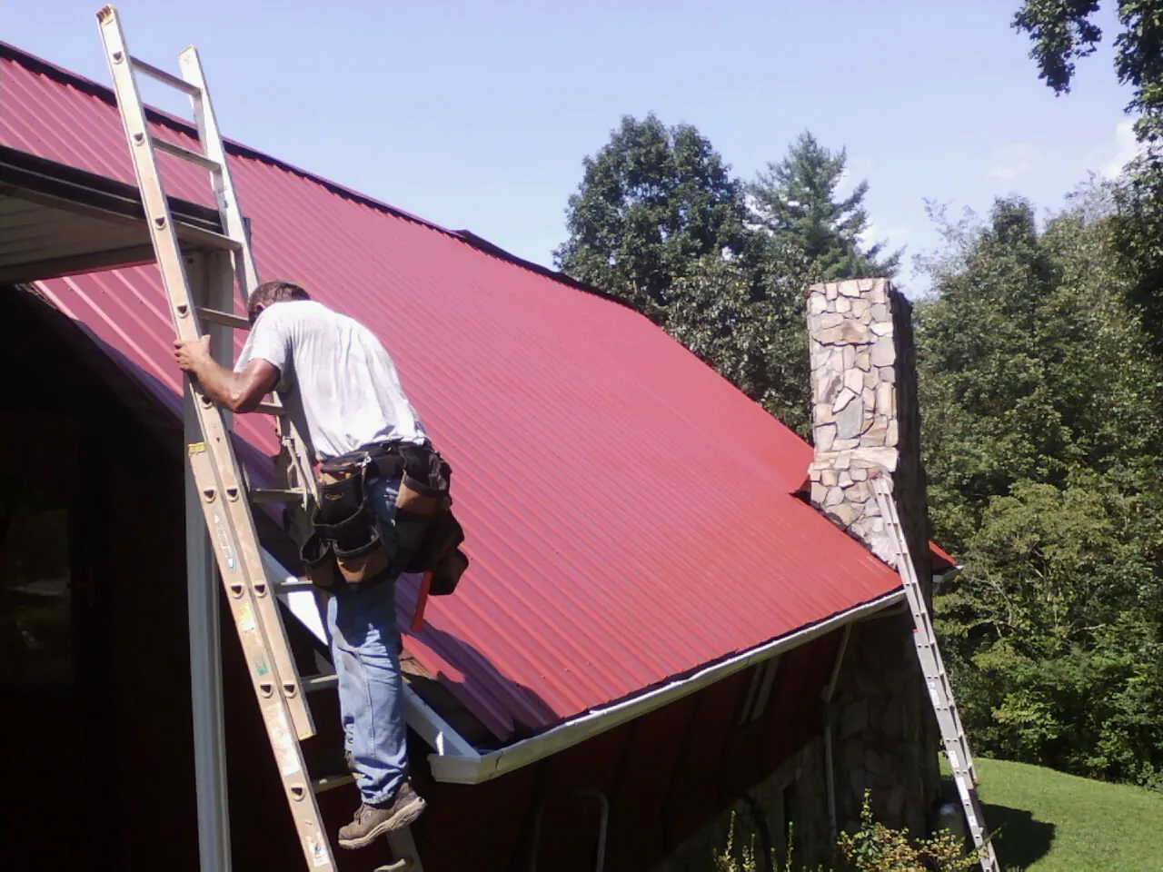 Roofer on a ladder beside a red metal roof and stone chimney — Soffit & Fascia Repair by Roofing Brothers in Ballwin